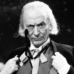 William Hartnell as The FIrst Doctor with his hands on his lapels and wearing a monocle